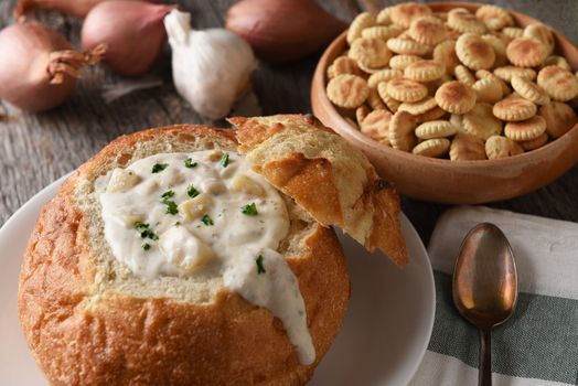 Closeup of a bread bowl of New England Clam Chowder. A bowl of oyster crackers, garlic and onions in the background.