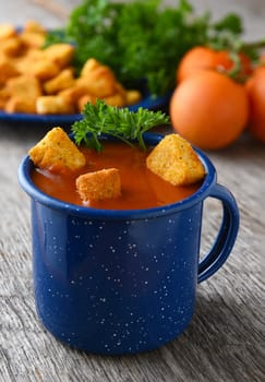 Closeup of a blue mug filled with fresh homemade tomato soup. Tomatoes, parsley and croutons out of focus in the background.