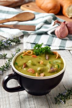 Split Pea Soup with ham and vegetables in a mug on a white rustic kitchen table with bread, towel, wood utensils and herbs.