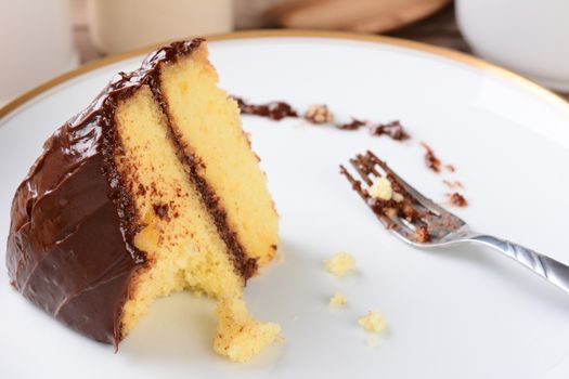 A slice of yellow cake with chocolate frosting with a piece missing. The white plate and fork have crumbs and frosting.