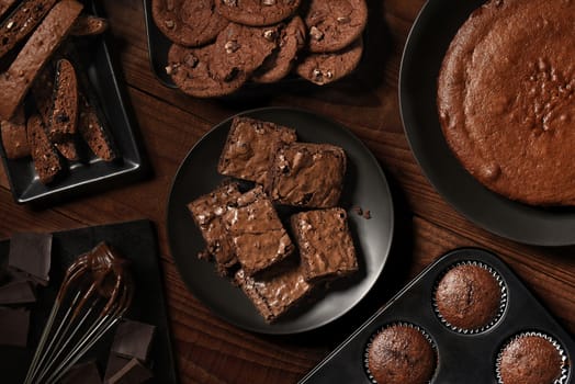Flat Lay display of fresh baked chocolate desserts on black plates and a rustic wood table. Items include, Biscotti, Cake, Muffins, Brownies, and Cookies.