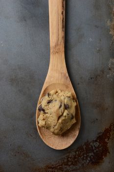 High angle closeup of a gob of chocolate chip cookie dough on a wooden spoon in the middle of an old used cookie baking sheet. Vertical format.