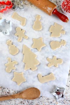 Christmas Cookie shapes on parchment paper. Around the perimeter are cutters, tub of sprinkles, spoon and rolling pin. Vertical format from a high angle.