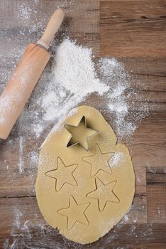 Top view of raw cookie dough with star shapes and cookie cutter on wood table with a rolling pin and flour sprinkles.