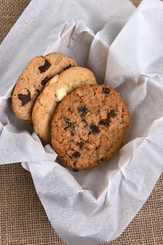 High angle view of a box of assorted gourmet cookies. Chocolate, oatmeal raisin and white chocolate chip cookies closeup in a box stuffed with paper on a burlap surface.