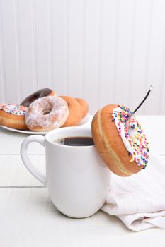 Closeup of a mug of hot coffee with a donut hanging off the spoon inside the cup. A plate of assorted donuts is in the background. Vertical with copy space.