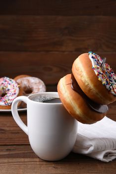 Closeup of a mug of hot coffee with two donuts hanging on the spoon inside the cup. A plate of assorted donuts is in the background. Vertical with copy space.