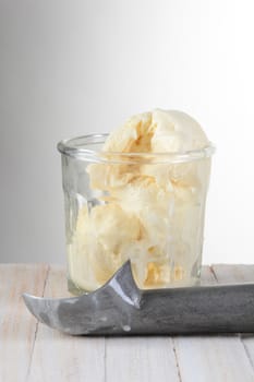 Glass of Vanilla Ice Cream and scoop on a wood table with a light to dark gray background. 