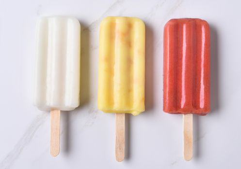 top view of three different ice pops on a marble counter top. Red, yellow and white fruit flavored pops.