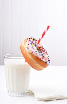 Closeup of a glass of milk with drinking straw going through the hole of a sprinkle covered donut. 