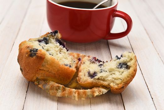 Closeup of a blueberry muffin with a cup of coffee in the background. The muffin is broken in half on a rustic white wood table.