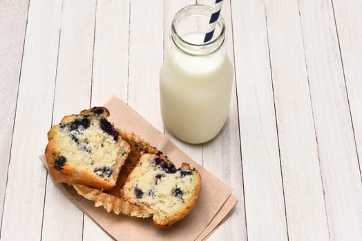 High angle view of a blueberry muffin and bottle of milk on a rustic white table. The muffin is broken in half on a napkin.