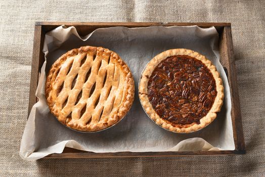 High angle view of an apple pie and pecan pie in a wood box lined with parchment paper. Horizontal format on a burlap table cloth.
