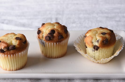 Closeup of three mini chocolate chip muffins on a white serving plate and a linen kitchen towel. 