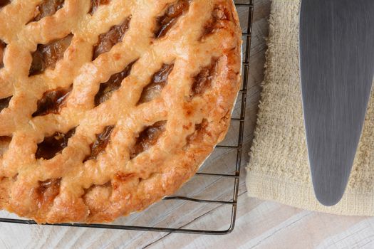 Closeup of a fresh baked apple pie on a cooling rack next to a serving spatula and towel. Horizontal format on a white rustic wooden kitchen table.