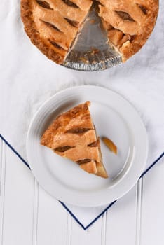 Overhead view of a slice of apple pie on a plate the rest of the pie is at the top of the frame. Vertical fomat on a white towel.