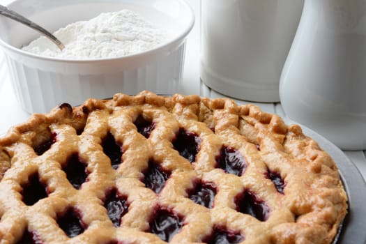 High angle shot of a fresh baked cherry pie with a lattice crust. A bowl of flour and pitchers fill the background. Horizontal format.