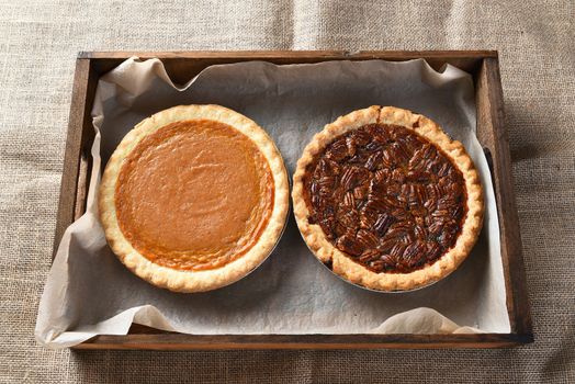 High angle view of a fresh baked pumpkin pie and a pecan pie in a wood box on burlap covered table, for Thanksgiving feast.