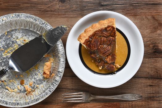 High angle view of a slice of pecan pie on a plate next to the empty pie tin with server. On a wood table with fork.
