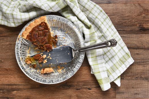 The last slice of a Thanksgiving holiday pecan pie. A tin with spatula on a wood table with kitchen towel. 