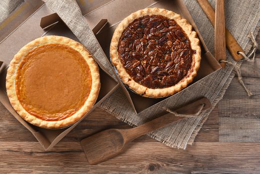 A pumpkin Pie and Pecan Pie in open bakery boxes on burlap and wood surface with wood utensils. 