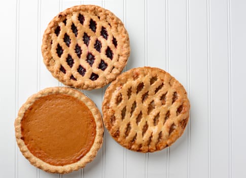 High angle shot of three pies, apple, pumpkin, and cherry. Horizontal format on a white beadboard surface. Thes pies are favorites for the  American Holidays, Thanksgiving and Christmas.