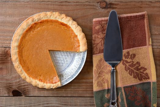 High angle shot of a pumpkin pie that has been cut into. Shot on a rustic wood table with a Autumn themed napkin and server by its side. Horizontal format.