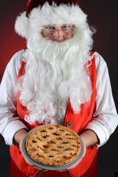 Closeup shot of Santa Claus serving a fresh baked Apple Pie. Santa is holding the dessert in both hands in front of his torso. Vertical format over a light to dark red background.