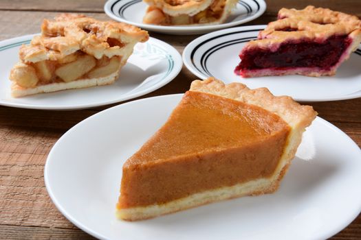 Closeup of four slices of pie on dessert plates. Focus is on the front slice of pumpkin pie. The back plates have apple and cherry pie. Horizontal format on an old wood kitchen table.