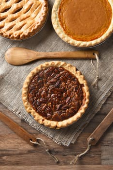 High angle vertical view of three pies for A thanksgiving feast. Pecan in the front with Apple and Pumpkin pies in the background. On burlap with wood utensils.