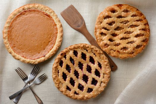 High angle shot of three fresh baked homemade pies, Apple, Cherry and Pumpkin on a burlap table cloth. Horizontal format with forks and wooden spatula.