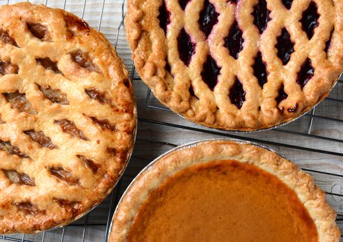 Three pies on cooling racks. High angle closeup shot of fresh baked apple, cherry and pumpkin pies on wire racks on a rustic wood kitchen table. Horizontal format.