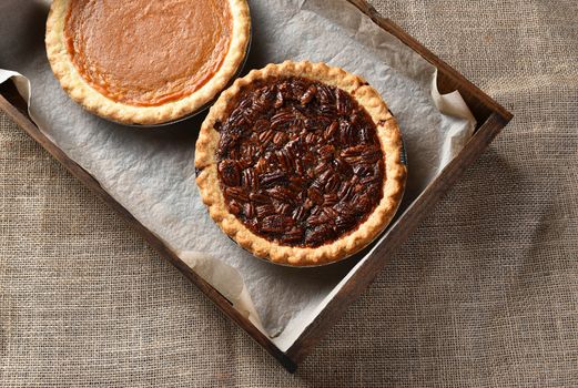 Top view of a piecan pie and pumpkin pie in a wood box on a burlap table cloth. 