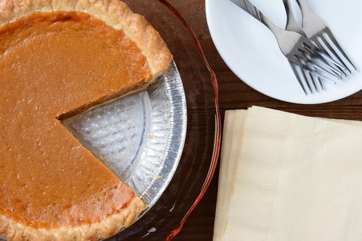 Top view of a fresh pumpkin pie with a slice missing with napkins, plates and forks.