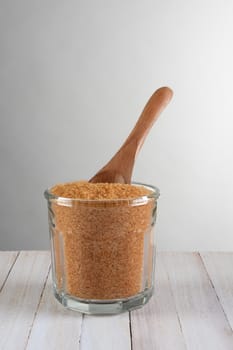 Closeup of a glass sugar bowl full of granulated brown sugar with a wooden spoon. On a rustic wood table against a light to dark gray background. Vertical format with copy space.