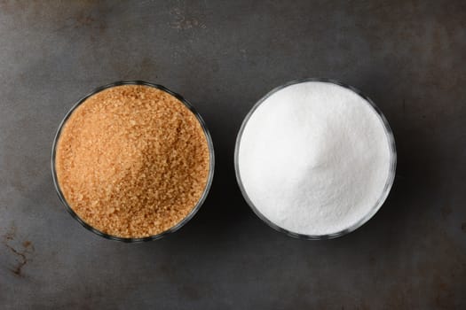 Two bowls of sugar. One bowl of granulated raw turbinado sugar and a second bowl of white granulated sugar. Horizontal format on a used baking sheet.