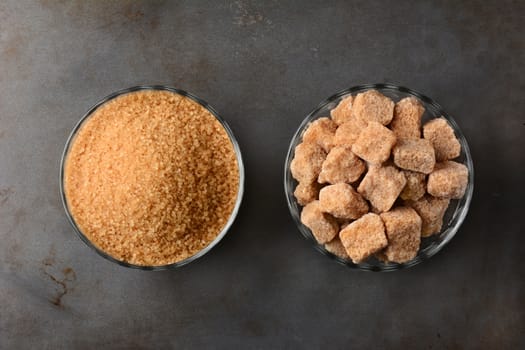 Two bowls of brown sugar. One bowl of granulated raw turbinado sugar and a second bowl of natural brown sugar lumps. High angle view on a used metal baking sheet