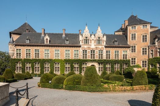 Gaasbeek, Flanders, Belgium - September 14, 2018: Southwest wing of Gaasbeek Castle with green garden in front, light brown walls, dark roof under blue sky.