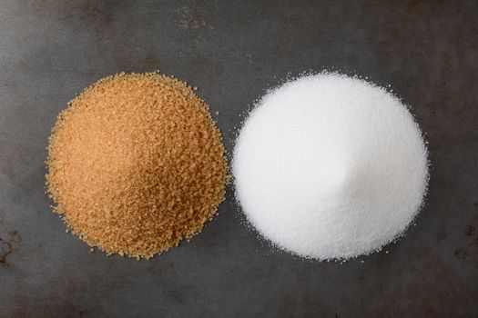 A pile of white granulated sugar and one of raw turbinado granules on a baking sheet. 