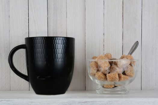 A black coffee mug and bowl of raw brown sugar cubes set in a rustic white wood kitchen. Horizontal format.