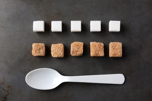 High angle view of white and brown sugar cubes and a white plastic spoon on a metal baking sheet. Five cubes of both the white and brown lumps line up in rows. Horizontal format.