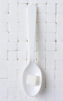 A white plastic spoon with a single sugar cube on a background of closely stacked white sugar cubes.