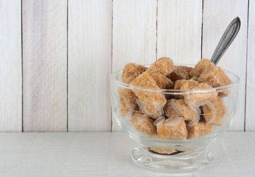 Closeup of a bowl full of raw brown sugar cubes. The bowl is on a white wood shelf against a rustic wood wall. Horizontal format with copy space.