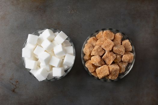 Bowls of sugar cubes. One bowl of raw brown sugar chunks and a second bowl of white sugar cubes. Horizontal format on a used baking sheet.