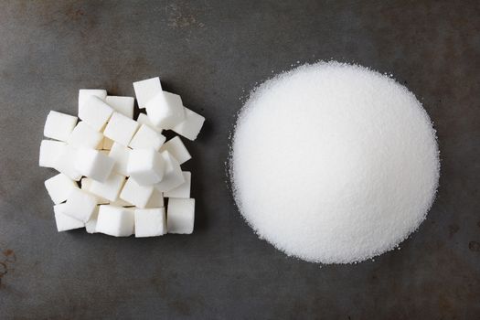 Overhead view of a pile of granulated white sugar and a mound of sugar cubes, on a used baking sheet.