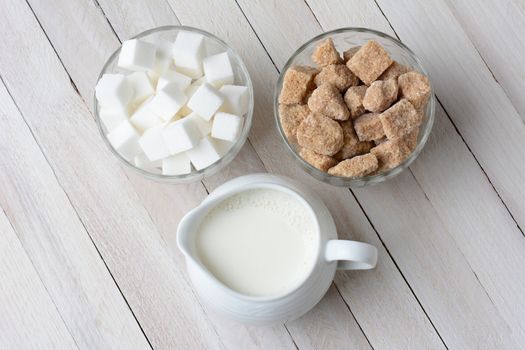 High angle shot of two bowls of sugar cubes, white and natural, and a pitcher filled with cream. Horizontal format on a rustic wood table.