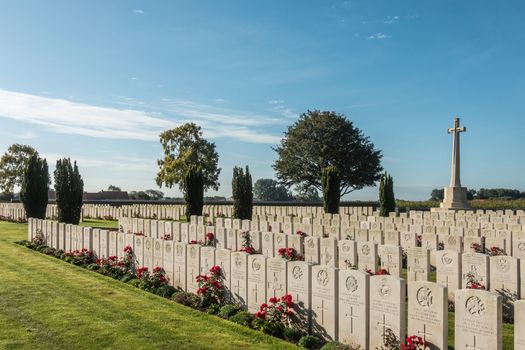 Proven, Flanders, Belgium - September 15, 2018: Overview of Mendinghem British war cemetery under blue morning sky. Green lawn, beige tomb stones and red roses with dark green trees sprinkled.