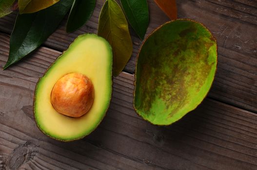 An Avocado cut in half on a wood surface with leaves. One half shows the seed while the other has been scooped out.