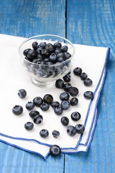 Closeup of a group of blueberries scattered on a napkin with a glass bowl filled with more berries. Vertical format on a rustic farmhouse style painted table, with shallow depth of field.