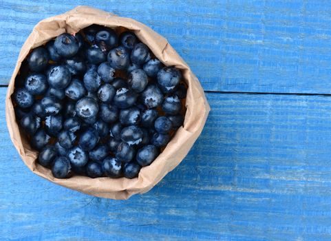 A brown paper bag full of fresh picked blueberries. The bag is on a blue wooden table.